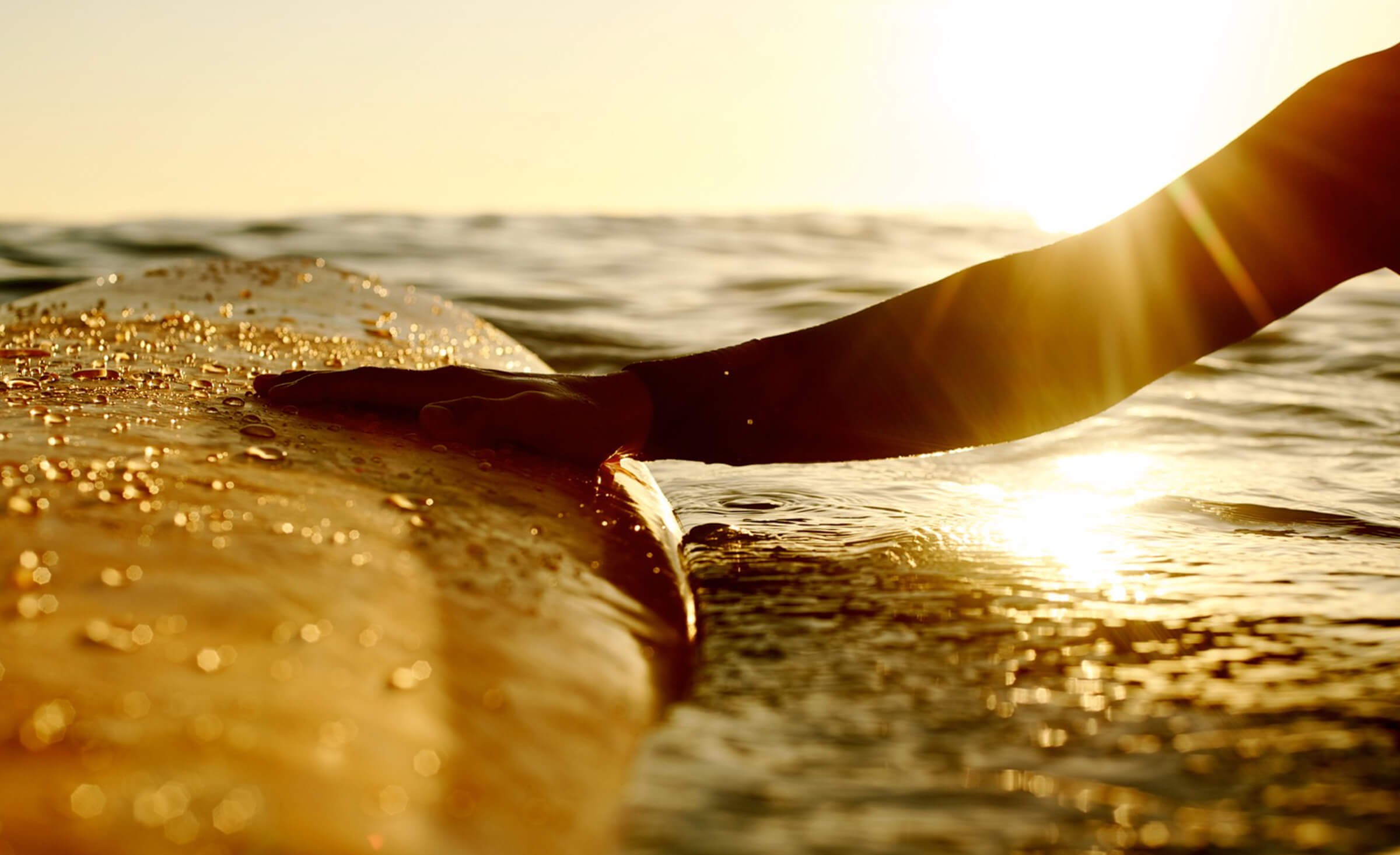 Hand reaches out to touch surfboard in ocean water with a rising sun in the background.